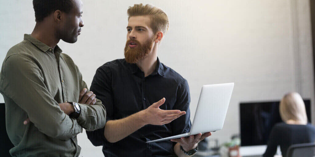 Two young smart business men discussing new project while standing in an office with laptop