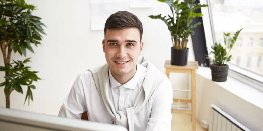 Horizontal portrait of attractive young brunette male architect sitting at his workplace in front of computer while working on new housing project using 3D CAD application, having happy expression