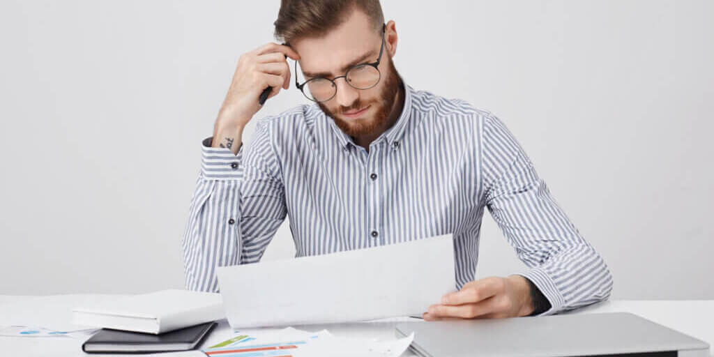 Serious concentrated bearded man dressed formally, reads document or contract, sits at work place, tries to understand figures and diagrams. Successful male enterpreneur studies office papers alone
