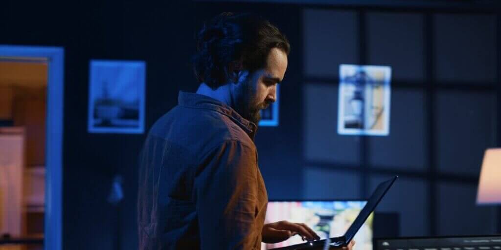 Man doing IT support job from home, walking around personal office with laptop in hand, coding. Nervous programmer wandering around room from stress, writing lines of code on notebook, camera A