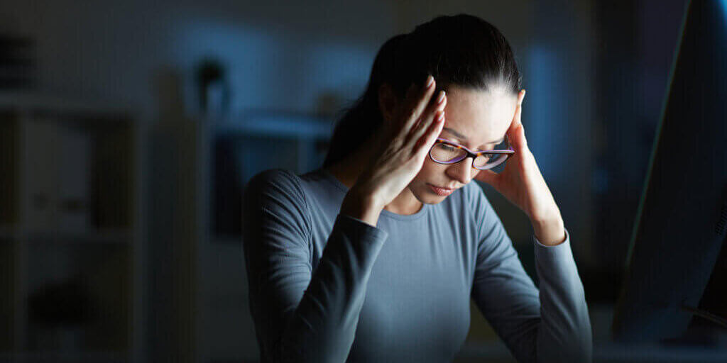 Young businesswoman with headache touching her head while sitting in front of computer monitor by her workplace at night